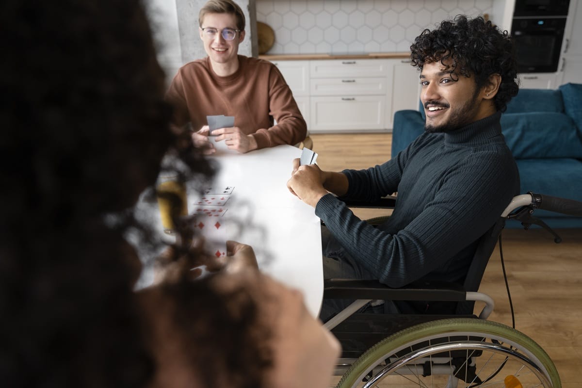 A smiling young man in a wheelchair playing a card game with friends at a table.