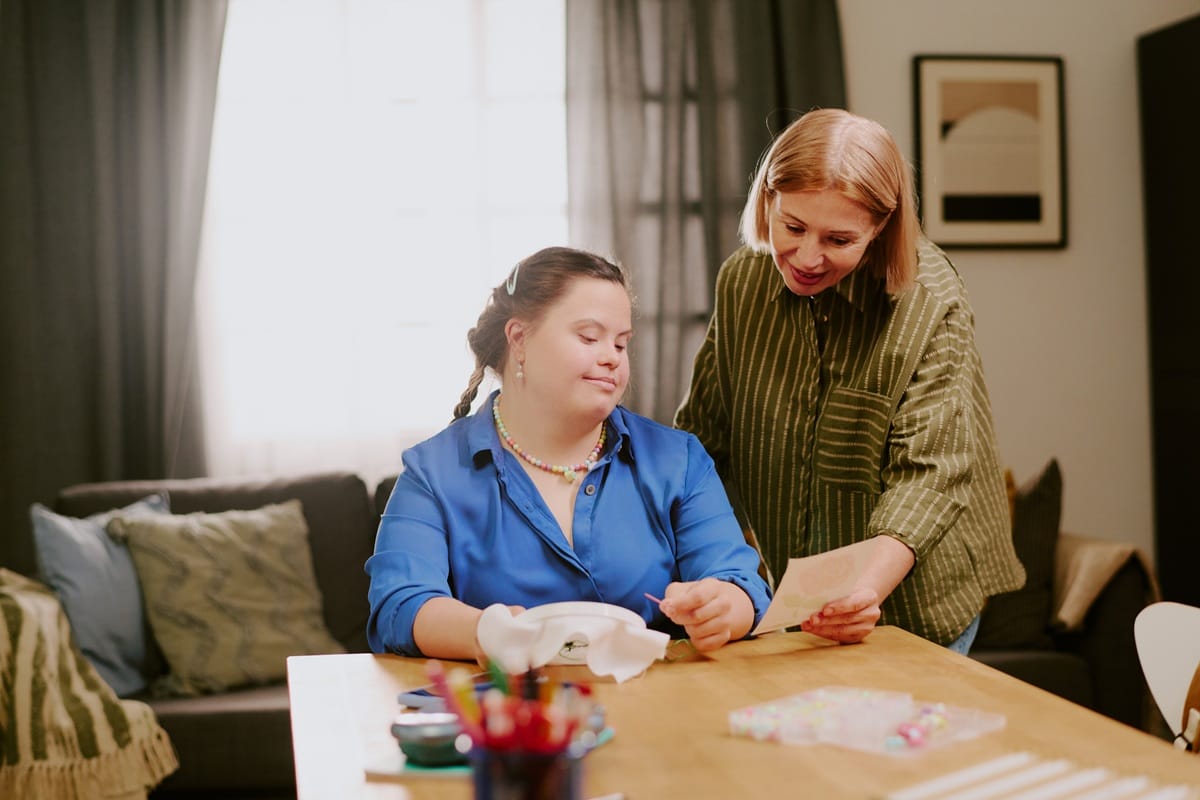 A support worker helps a seated young woman with an embroidery craft at a table, highlighting tailored routines