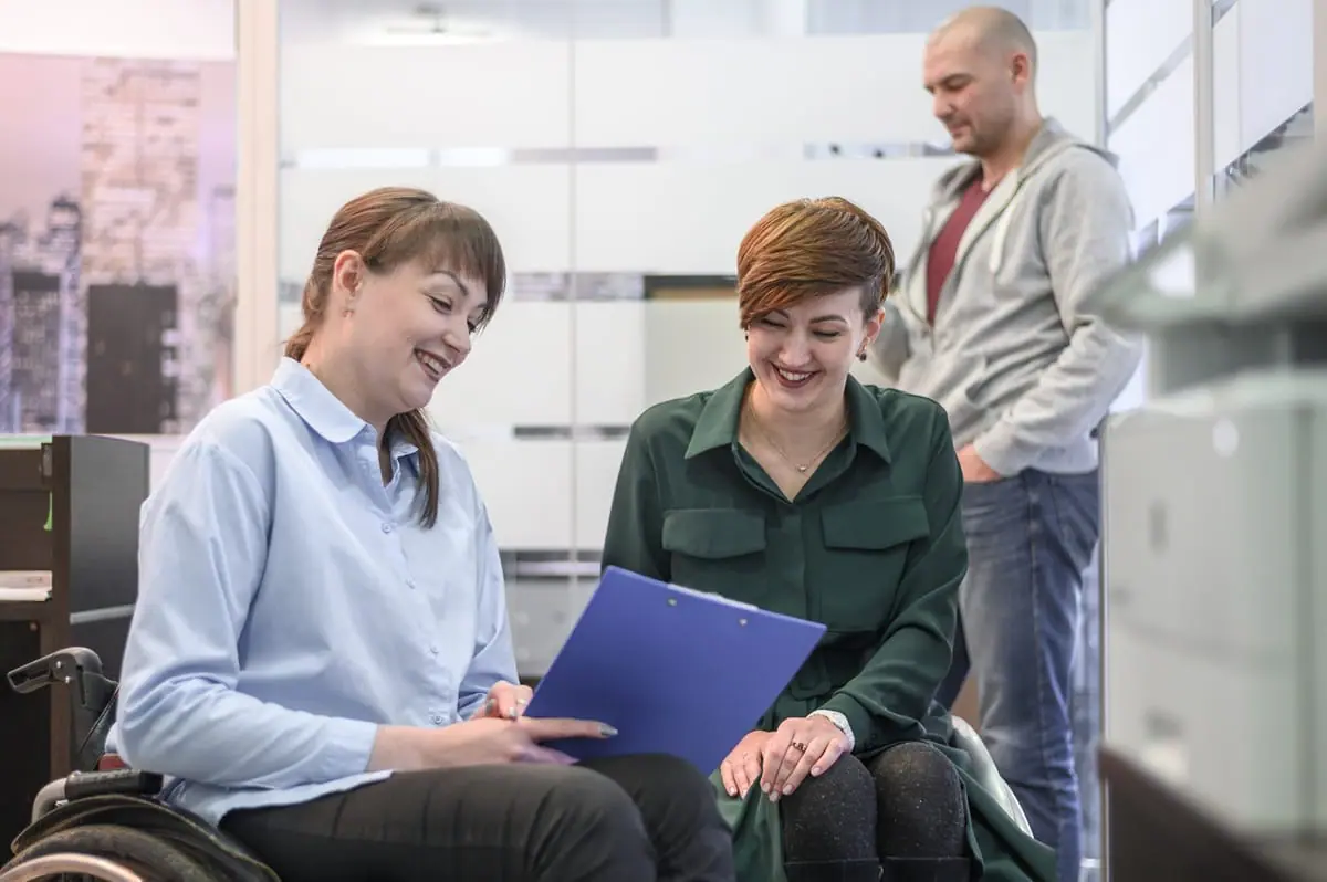 A woman in a wheelchair smiling while reviewing NDIS application documents with a support worker in an office setting.