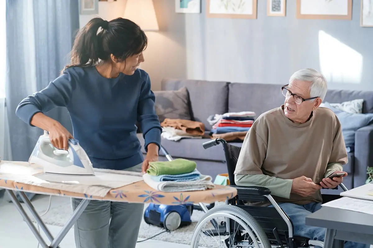 A support worker irons clothes while conversing with an older man in a wheelchair holding a smartphone