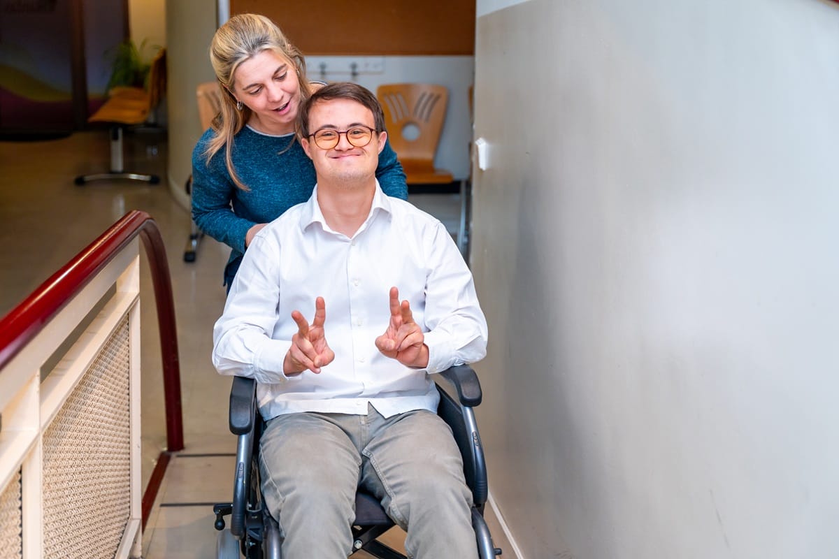 A smiling support worker pushes a young man wearing glasses in a manual wheelchair down a hallway