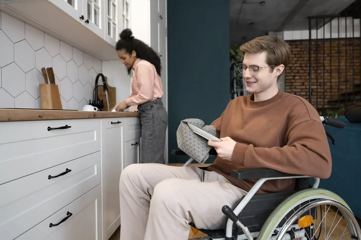 A smiling man in a wheelchair uses a tablet in a kitchen while someone assists in the background, highlighting choice and control.
