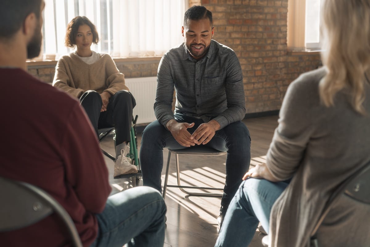 A smiling man speaks to a diverse NDIS support group, including a wheelchair user, seated in a circle