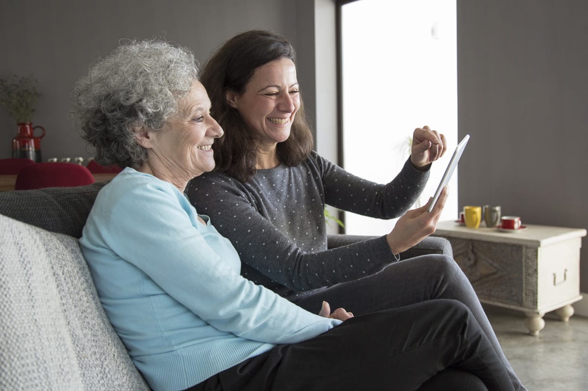 Two smiling women sitting on a couch reviewing information on a tablet, helping explain care funding and budgets.