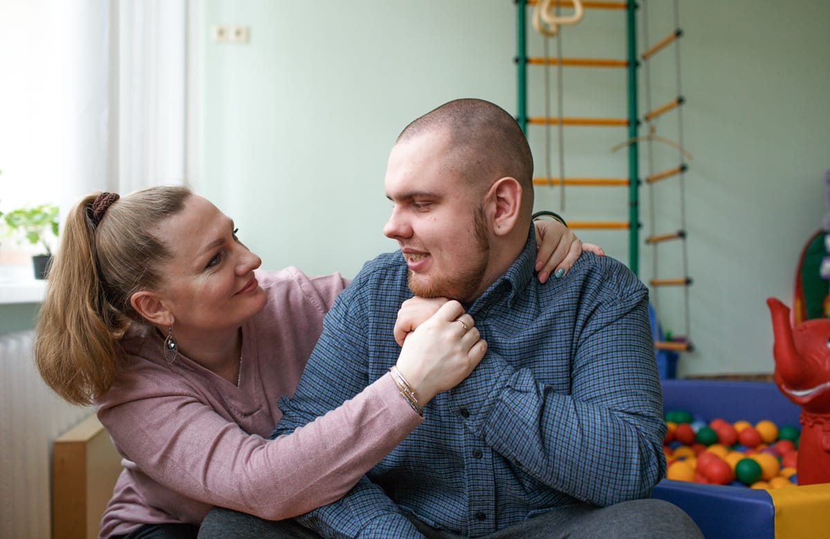 A woman and a young man smile warmly at each other in a room with therapy equipment, representing supportive living arrangements.