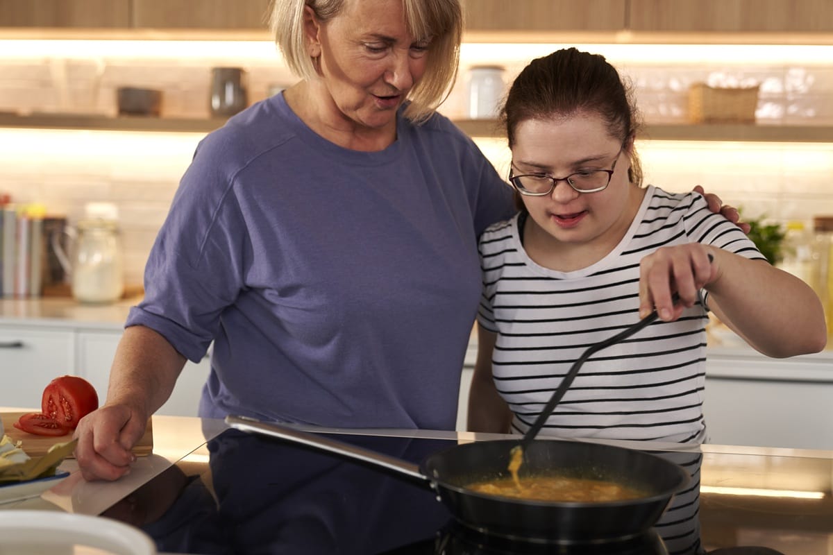 A young woman with Down syndrome cooks at a stove with the support of an older woman, practising daily living skills like meal preparation.