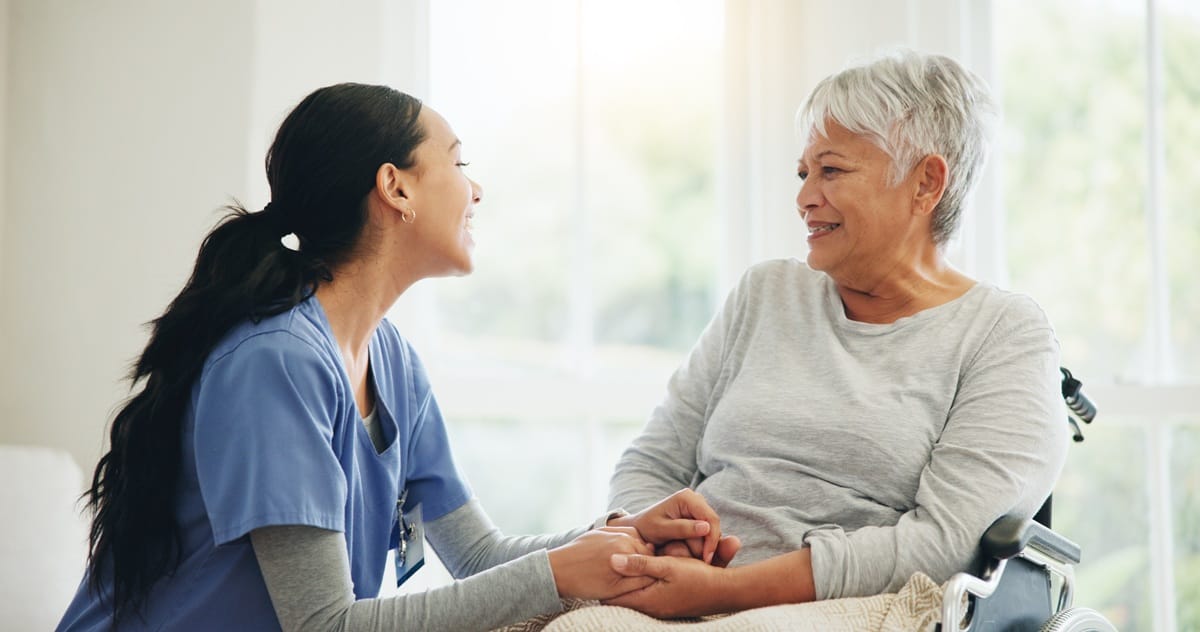 A smiling caregiver in blue scrubs holding hands with an older woman, representing compassionate Support at Home services.