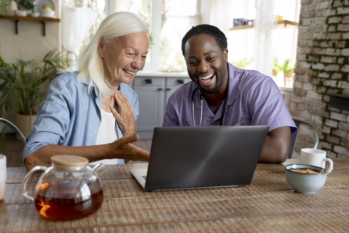 A smiling male healthcare worker and an older woman looking at a laptop at a table, discussing care program options.