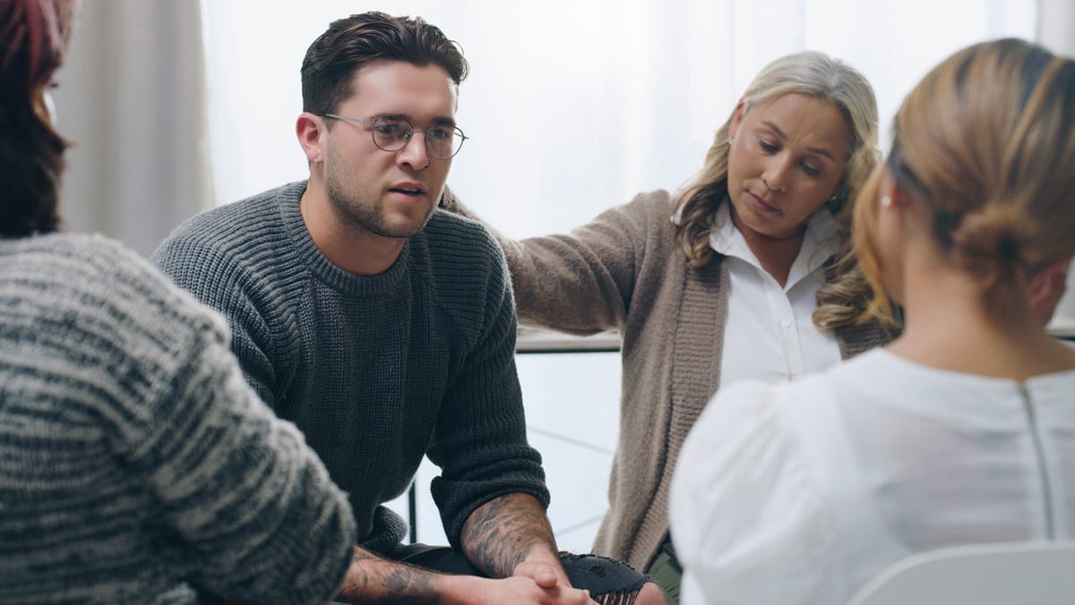 A young man speaks earnestly in a support circle while a woman rests a comforting hand on his shoulder