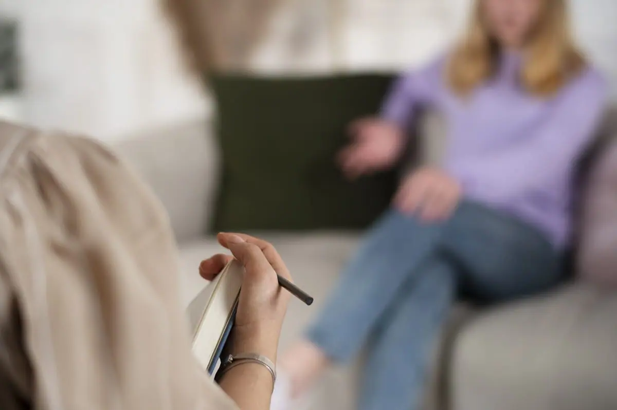 Over-the-shoulder view of a recovery coach holding a notebook, listening to a woman talking on a couch