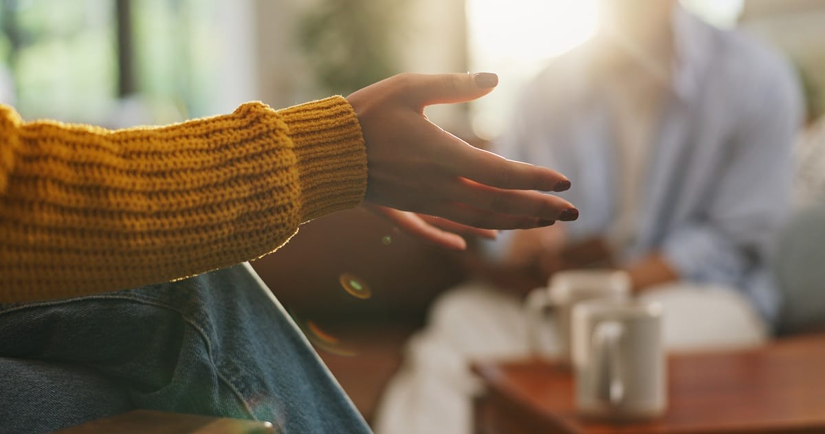 Close-up of a person in a yellow sweater gesturing warmly over coffee, representing safe psychosocial coaching support