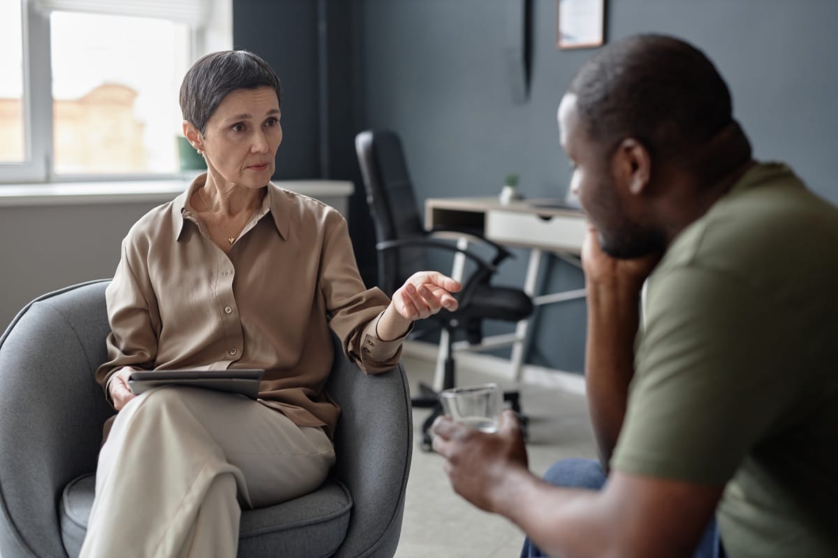 An older woman in a tan shirt gesturing while speaking with a man, illustrating a consultation for NDIS support services.