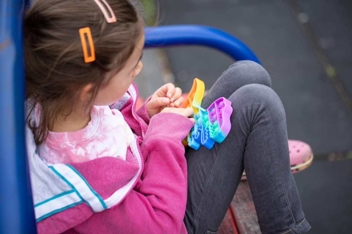 Young girl sitting on playground equipment playing with a rainbow-colored pop-it fidget toy to support sensory regulation.