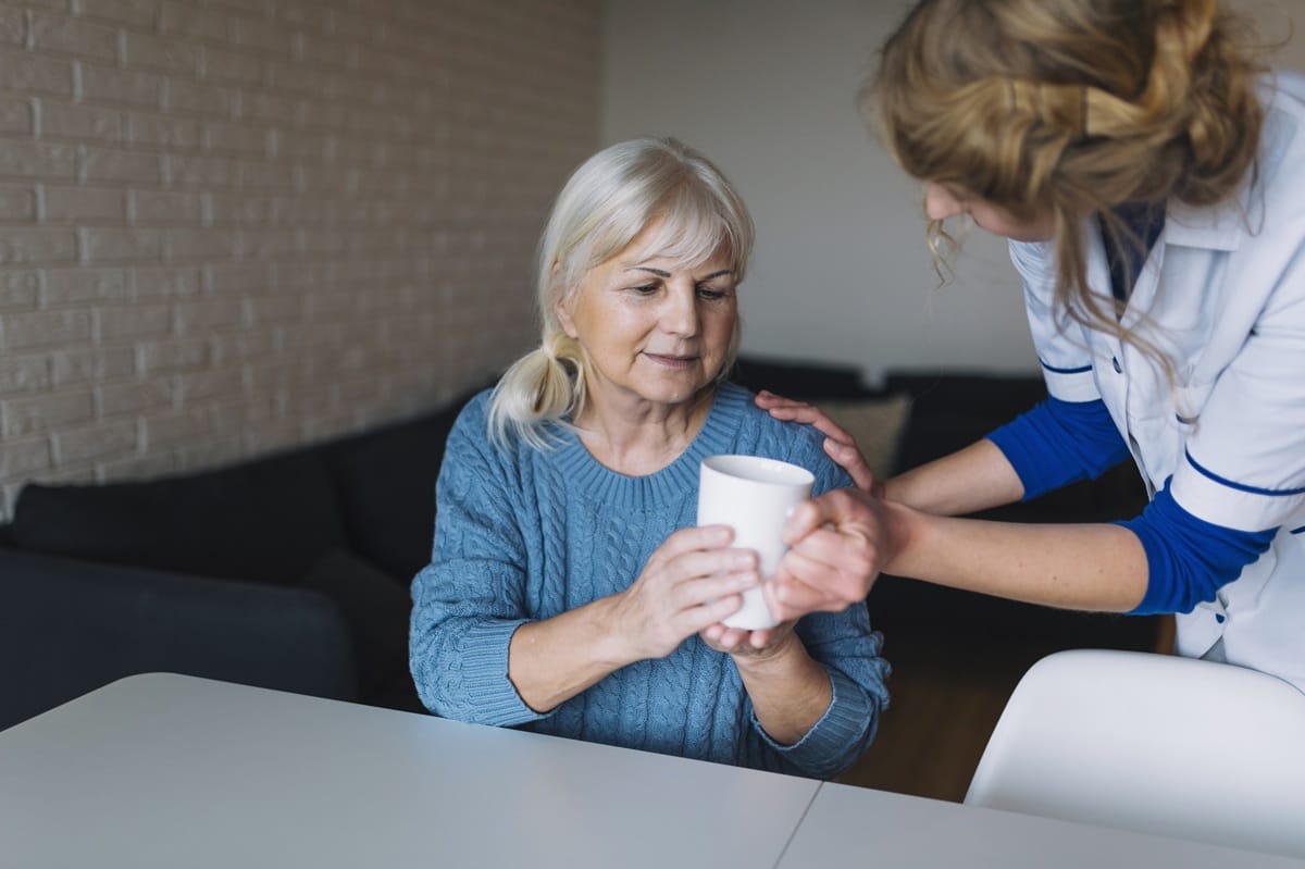 Support worker in a white uniform handing a white mug to an older woman sitting at a home dining table