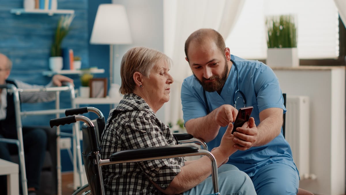 Support worker in blue uniform and an older woman in a wheelchair looking at a smartphone together