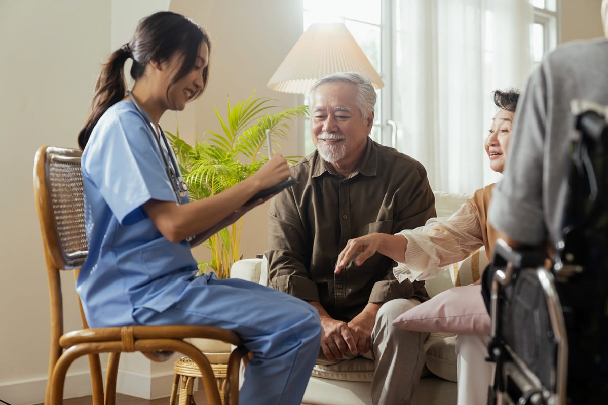 Support worker in blue scrubs writing on a clipboard while smiling at an older couple sitting on a sofa