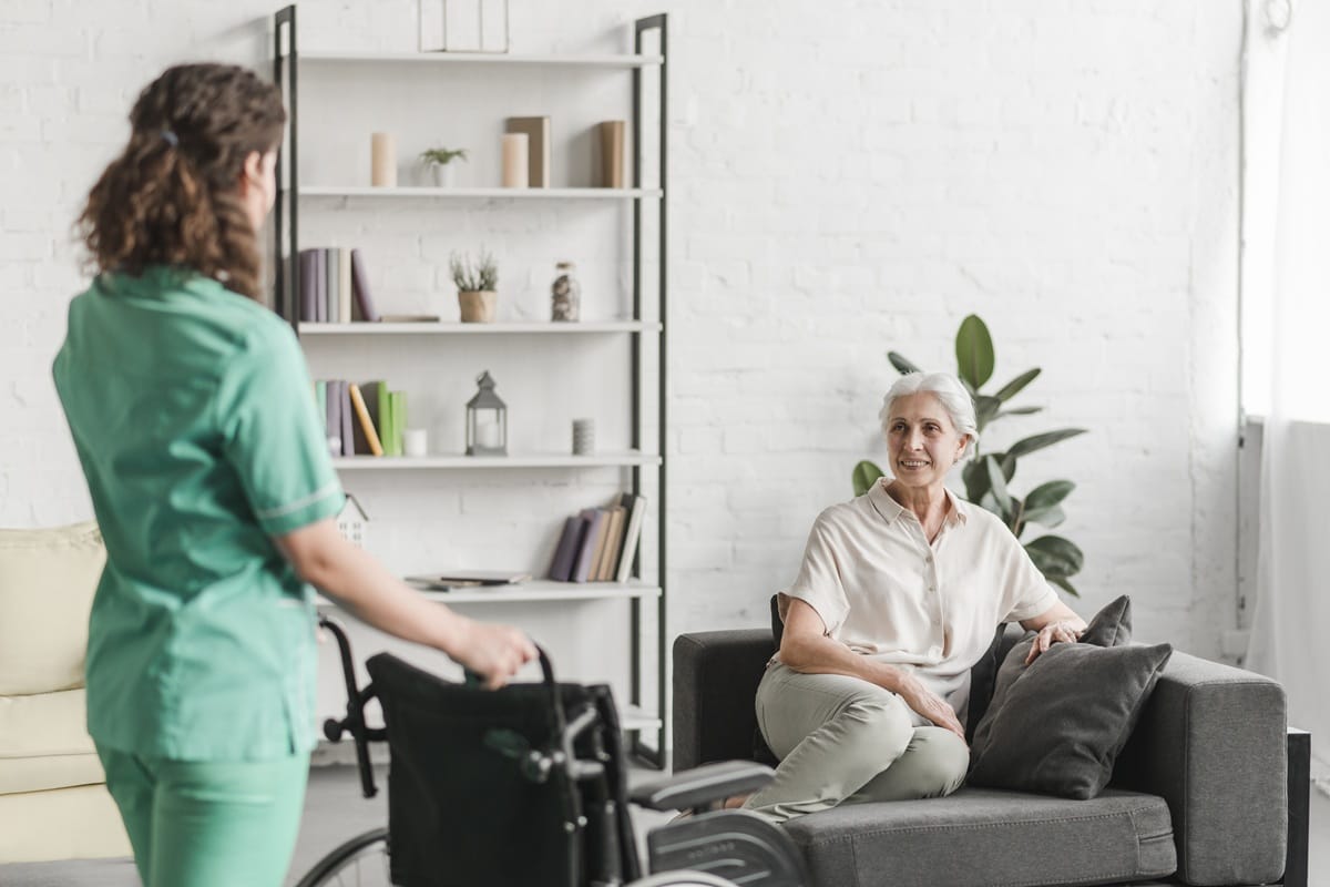 Support worker in green uniform with a wheelchair approaching a smiling older woman sitting on a couch